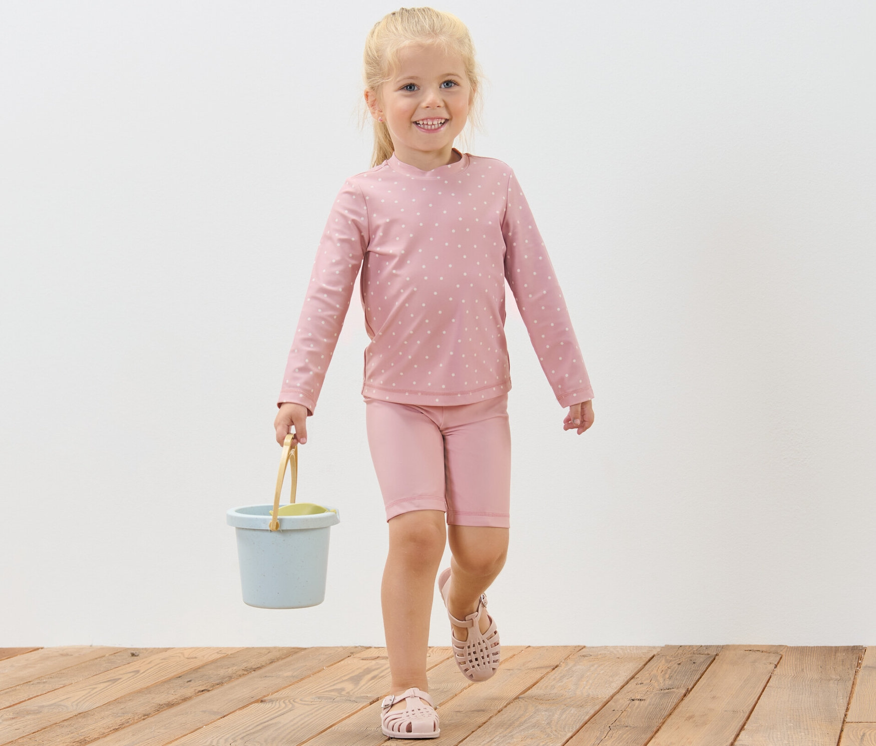 Ein lächelndes Mädchen geht auf einem Holzboden und trägt einen blauen Eimer. Sie trägt eine Kinder-Kombi mit UV-Schutzfaktor 80, Punkte.