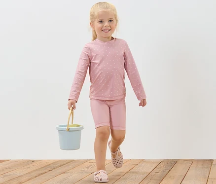 Ein lächelndes Mädchen geht auf einem Holzboden und trägt einen blauen Eimer. Sie trägt eine Kinder-Kombi mit UV-Schutzfaktor 80, Punkte.