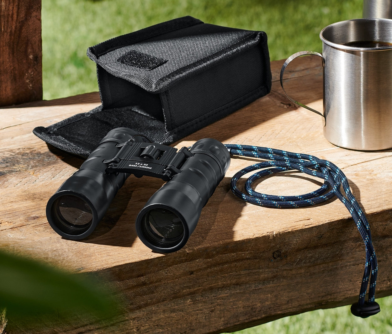 Auf einer Holzbank liegen ein Taschenfernglas »12x32«, ein Etui, ein Metallbecher und eine Flasche.