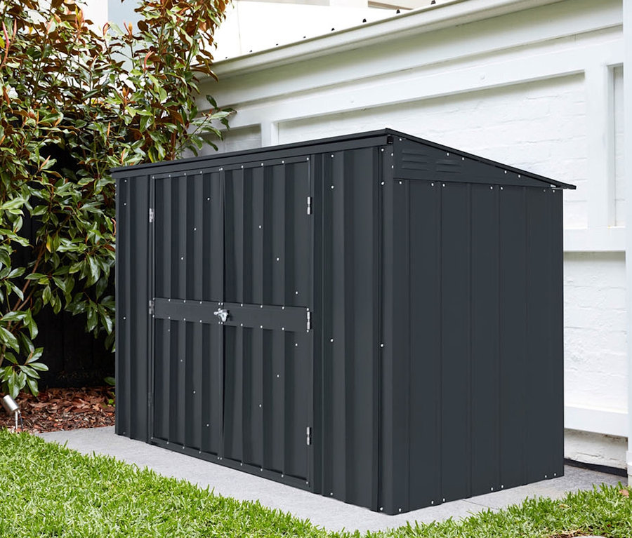 Eine graue Gerätebox steht auf einer Betonfläche neben einer weißen Wand und einem grünen Busch.