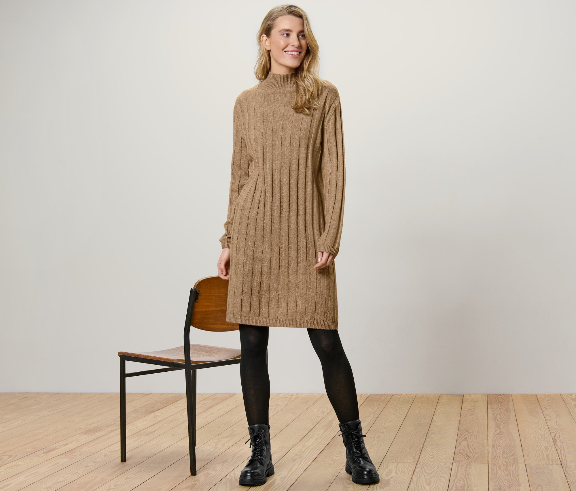 Eine Frau steht neben einem Stuhl und trägt ein geripptes Strickkleid mit Stehkragen.