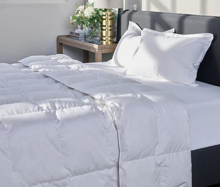 Ein gemachtes Bett mit grauer Kopflehne, zwei weißen Kissen und einer weißen, gesteppten Home By TEMPUR® kuschelige Luxe Duo-Decke. Auf dem Nachttisch stehen Lampe und Blumen.