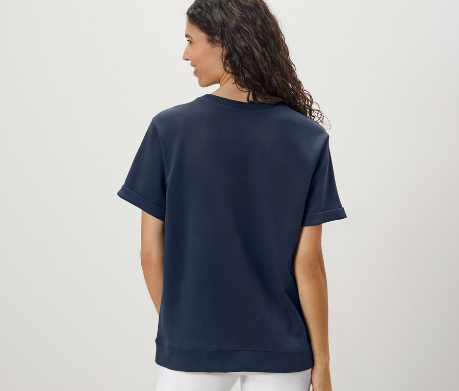 Rückansicht einer Frau mit gewelltem Haar, die ein dunkelblaues Shirt aus Modalmix und eine weiße Hose trägt.