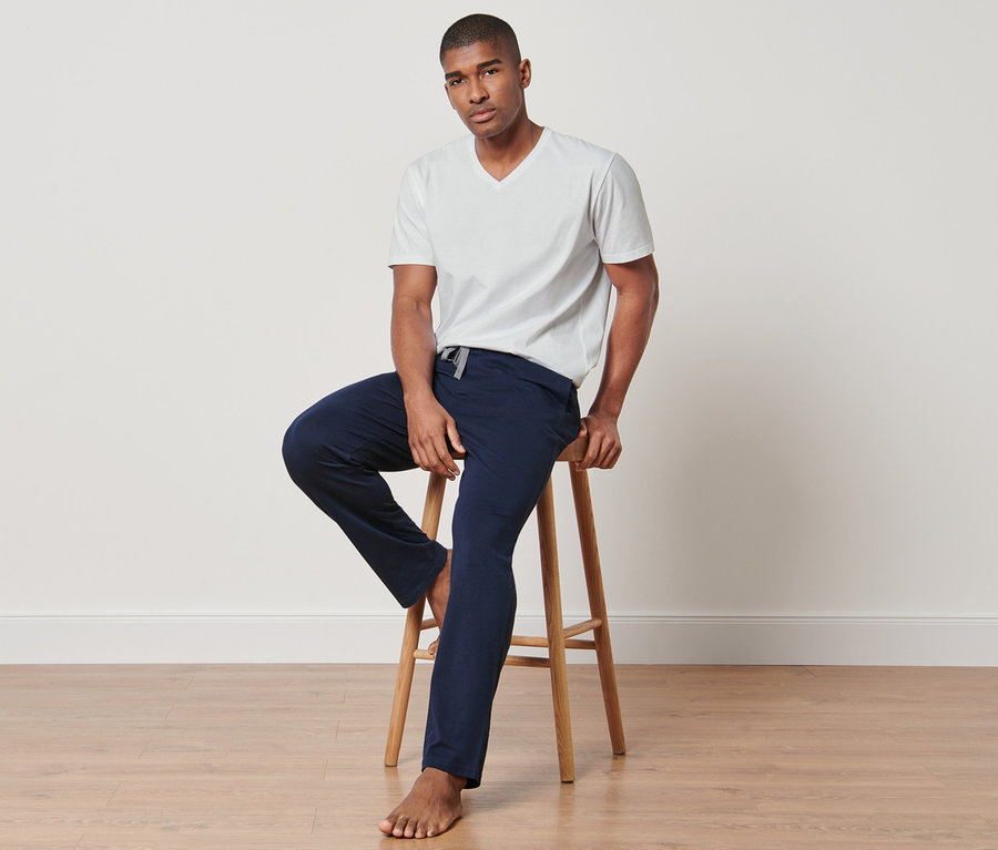 Mann sitzt auf einem Hocker und trägt ein weißes T-Shirt und eine dunkelblaue Hose aus einem 4-teiligen Pyjama-Set.