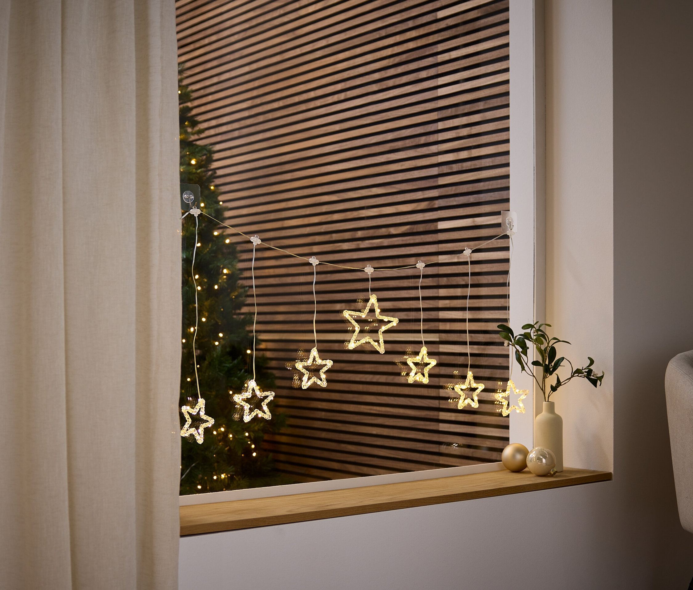 Lichterkette in Sternform hängt am Fenster, dahinter ist ein Weihnachtsbaum zu sehen.