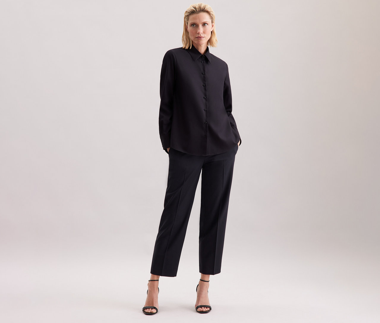 Eine blonde Frau steht in einer schwarzen Seidensticker Regular-Bluse, einer schwarzen Hose und schwarzen Sandalen vor einem hellen Hintergrund.