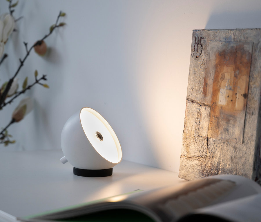 Eine weiße Lampe steht auf einem weißen Tisch neben einem offenen Buch und einem Bild.