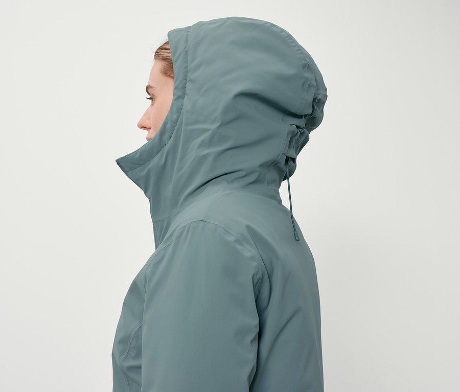 Profil einer Frau mit Kapuze in einer blaugrünen Jacke vor einem weißen Hintergrund.