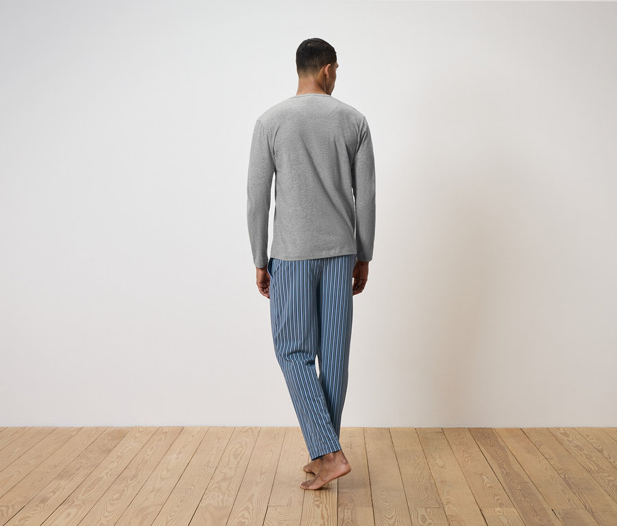 Ein Mann mit dem Rücken zur Kamera steht auf einem Holzboden. Er trägt ein graues Langarmshirt und eine blau gestreifte Pyjamahose.