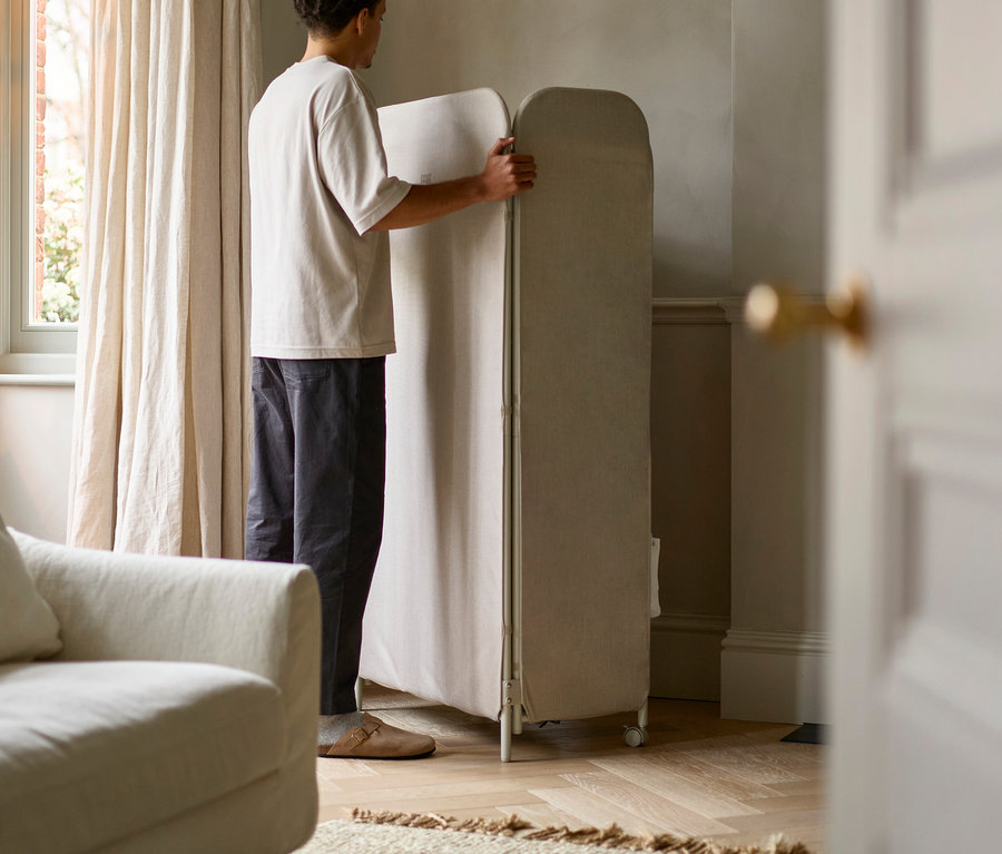Mann hantiert mit einer beige-farbenen, zusammenklappbaren Sichtschutzwand mit Rollen im Wohnzimmer.