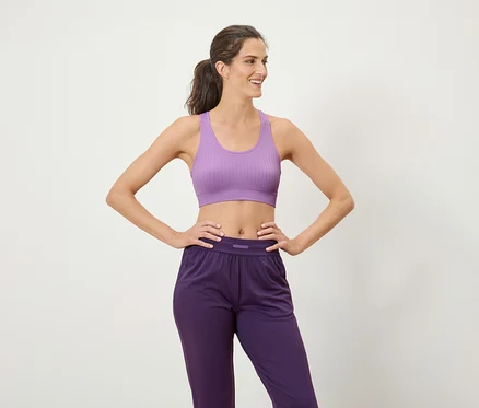 Frau mit braunen Haaren im Pferdeschwanz posiert in einem lila Sport-BH und einer violetten Sporthose.