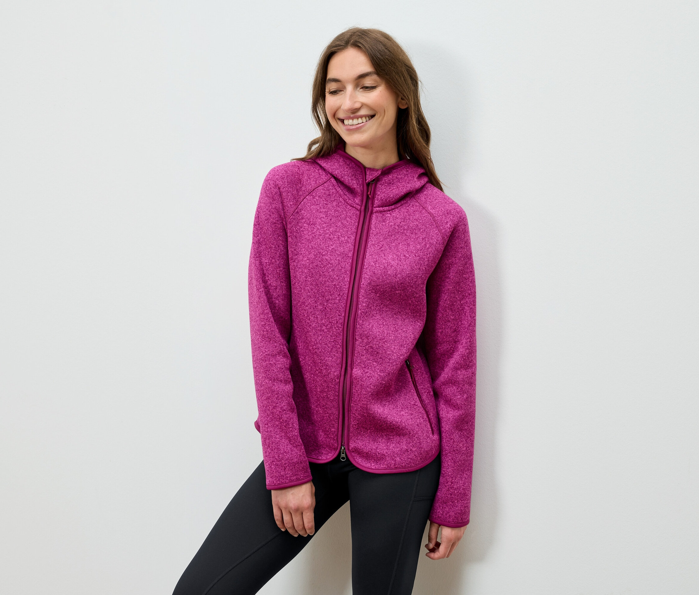 Lächelnde Frau posiert in einem pinkfarbenen Kapuzenpullover und schwarzen Hosen.