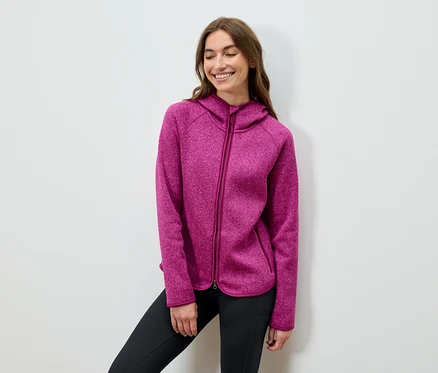Lächelnde Frau posiert in einem pinkfarbenen Kapuzenpullover und schwarzen Hosen.