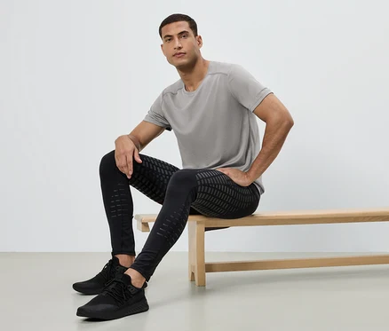 Mann sitzt auf einer Bank, trägt ein graues T-Shirt, schwarze Leggings mit Muster und schwarze Turnschuhe.