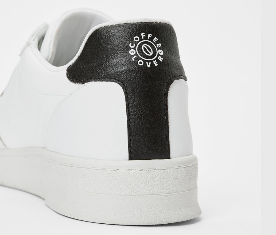Detailaufnahme eines weißen Tchibo x VLACE Coffee Lover Sneakers mit schwarzem Rand und Coffee Lover Logo.