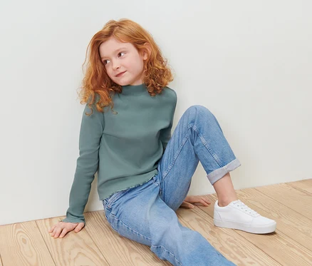 Ein rothaariges Mädchen sitzt auf einem Holzboden in einem grünen Langarmshirt und blauen Jeans.