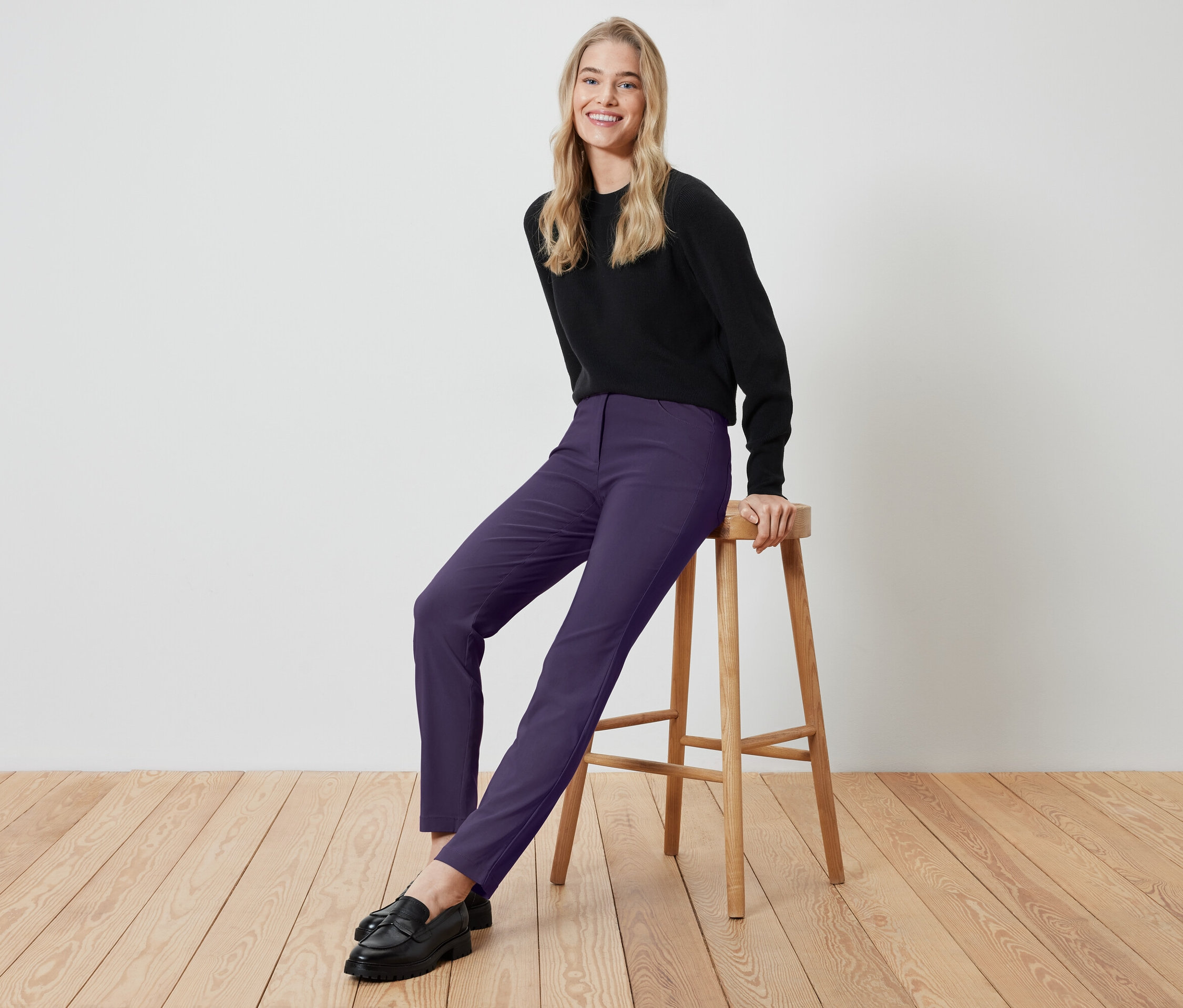Eine blonde Frau sitzt auf einem Holzhocker und trägt ein schwarzes Oberteil und violette Hosen.