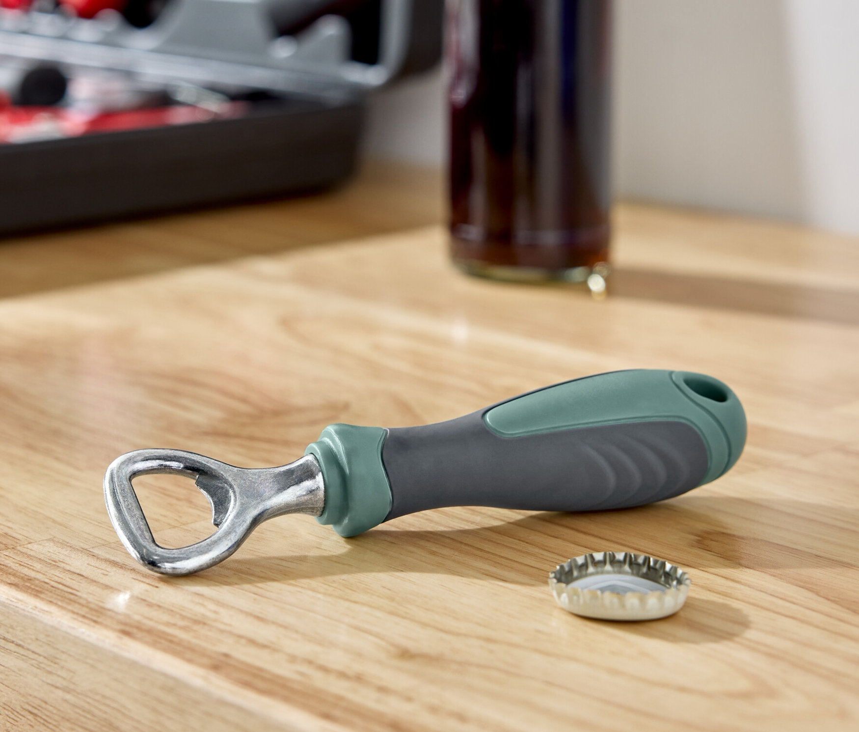 Auf einer Holzfläche liegen ein Flaschenöffner neben einem Kronkorken, im Hintergrund eine Flasche und ein Werkzeugkasten.