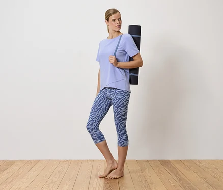 Frau mit blonden Haaren trägt eine Yogamatte über der Schulter. Sie trägt ein lila T-Shirt und blaue gemusterte Leggings.