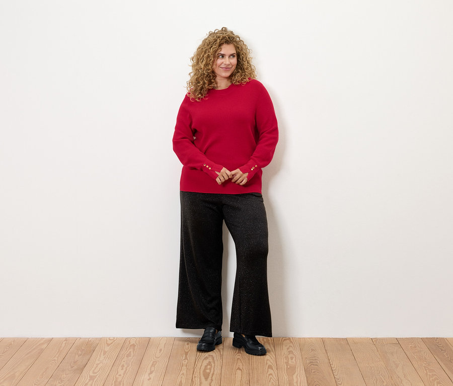 Frau mit lockigem Haar steht und trägt einen roten Pullover und eine schwarze Hose.