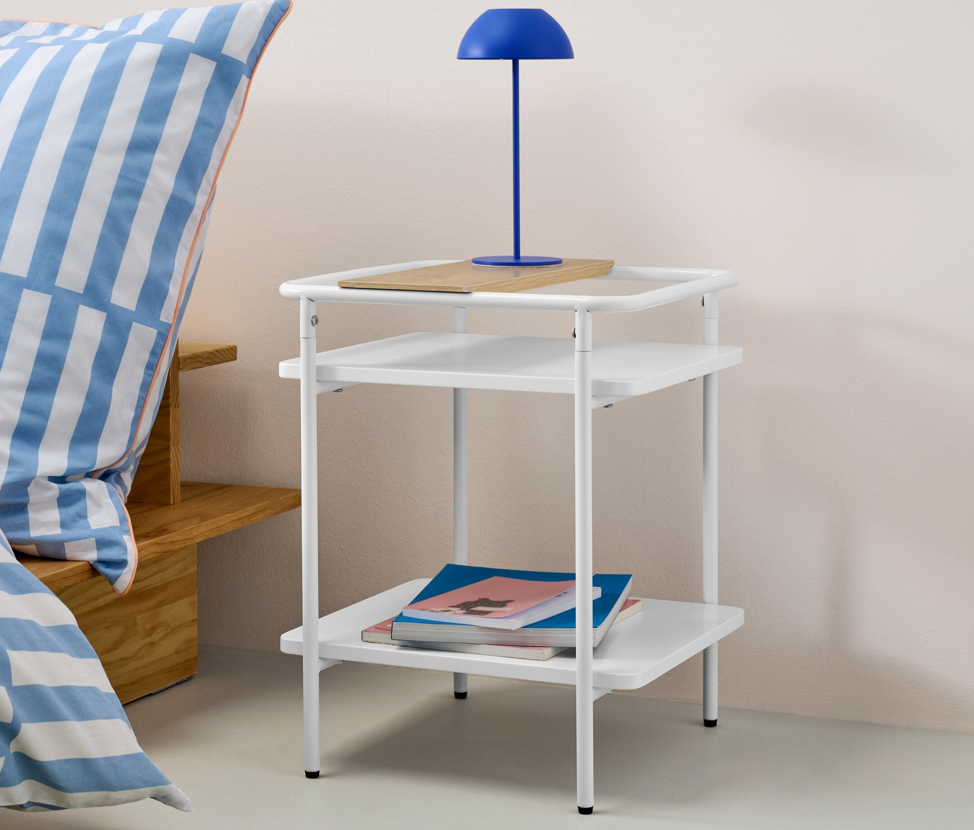 Weißer Beistelltisch mit zwei Ablagen, auf der oberen Ablage ein Holzbrett und darauf eine blaue Lampe. Auf der unteren Ablage liegen Bücher.