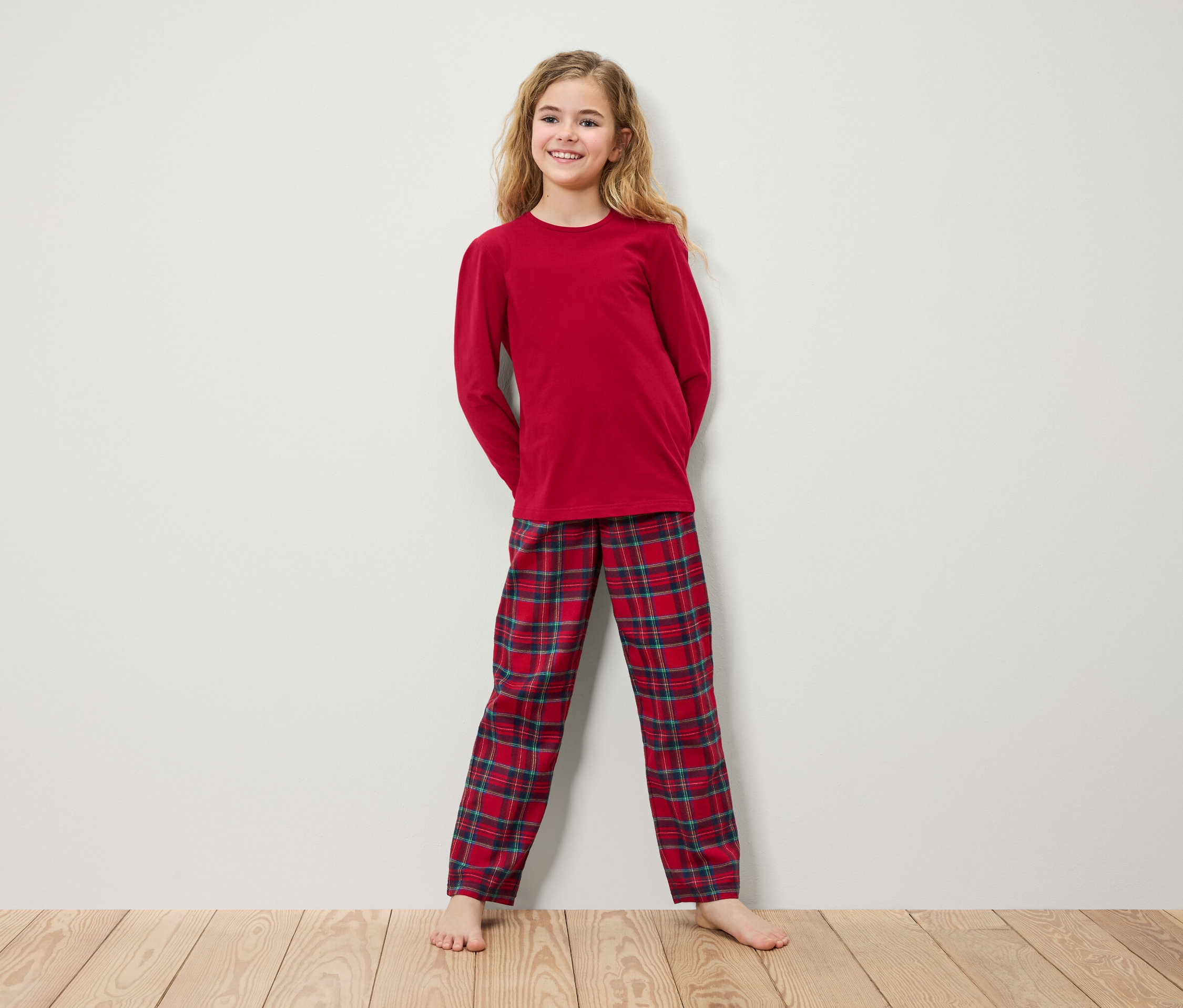 Mädchen im roten Langarm-Pyjama und karierter Hose steht auf einem Holzboden vor einer weißen Wand.