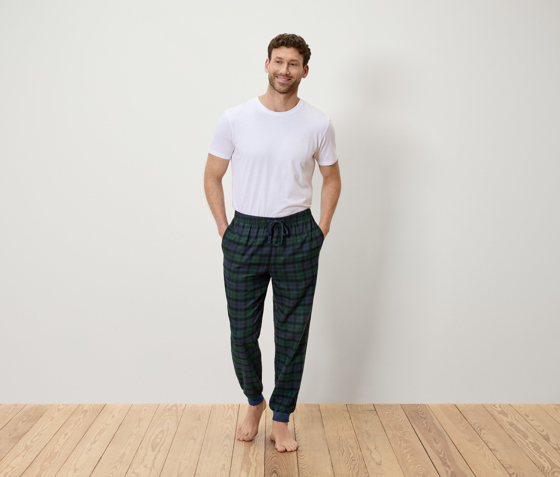 Mann steht mit den Händen in den Hosentaschen und trägt ein weißes T-Shirt und eine grün-blau karierte Hose.