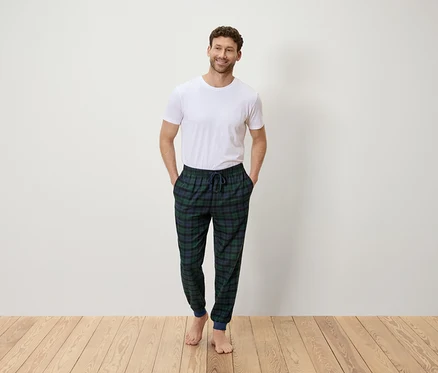 Mann steht mit den Händen in den Hosentaschen und trägt ein weißes T-Shirt und eine grün-blau karierte Hose.