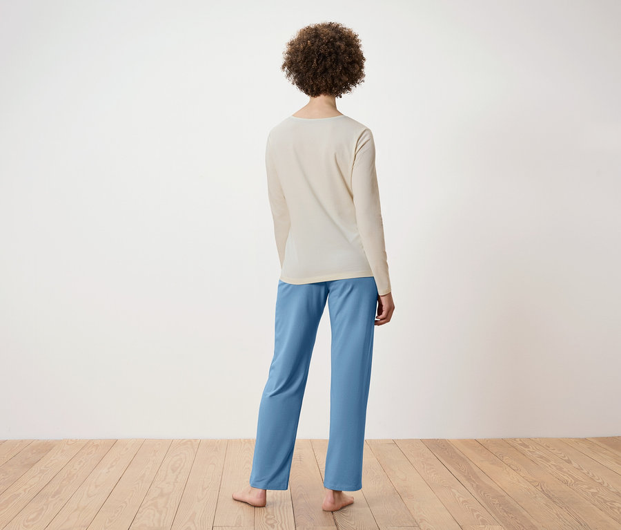 Von hinten ist eine Frau mit lockigem Haar zu sehen, die ein beiges Langarmshirt und eine blaue Hose trägt. Sie steht auf einem Holzboden.