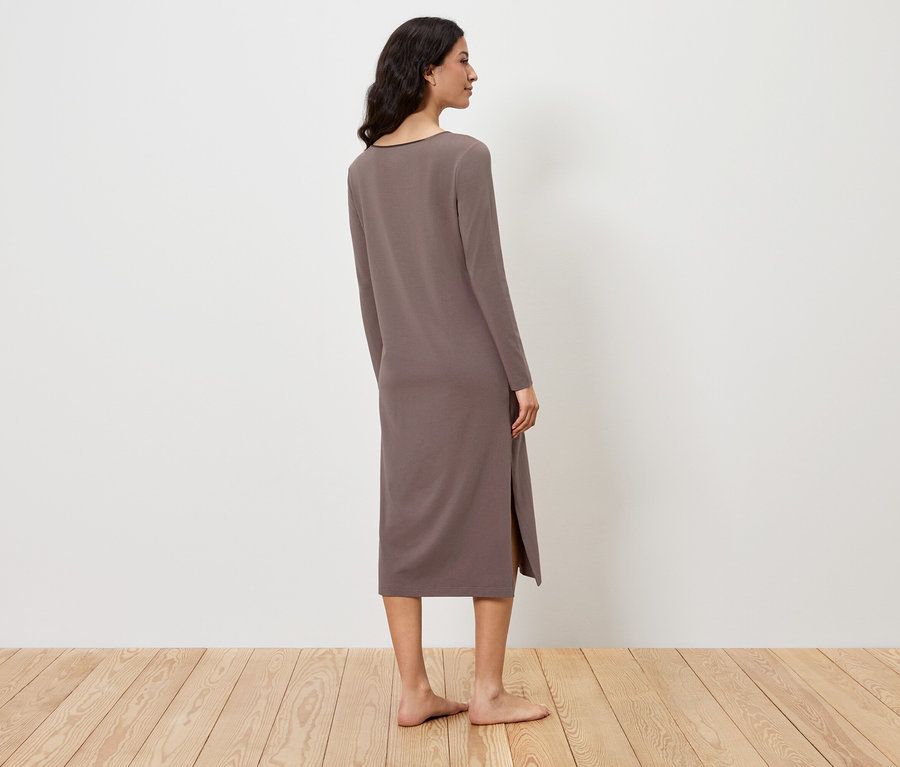 Eine Frau steht mit dem Rücken zum Betrachter und trägt ein langes, braunes Kleid mit langen Ärmeln und Schlitz.