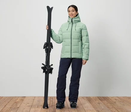 Frau hält Ski und trägt eine grüne Jacke und dunkelblaue Hosen.