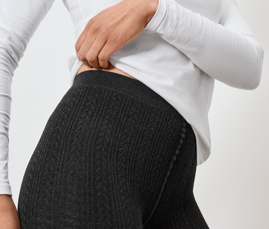 Frau in grauen Strickleggings und weißem Langarmshirt, die den Bund der Leggings anpasst.