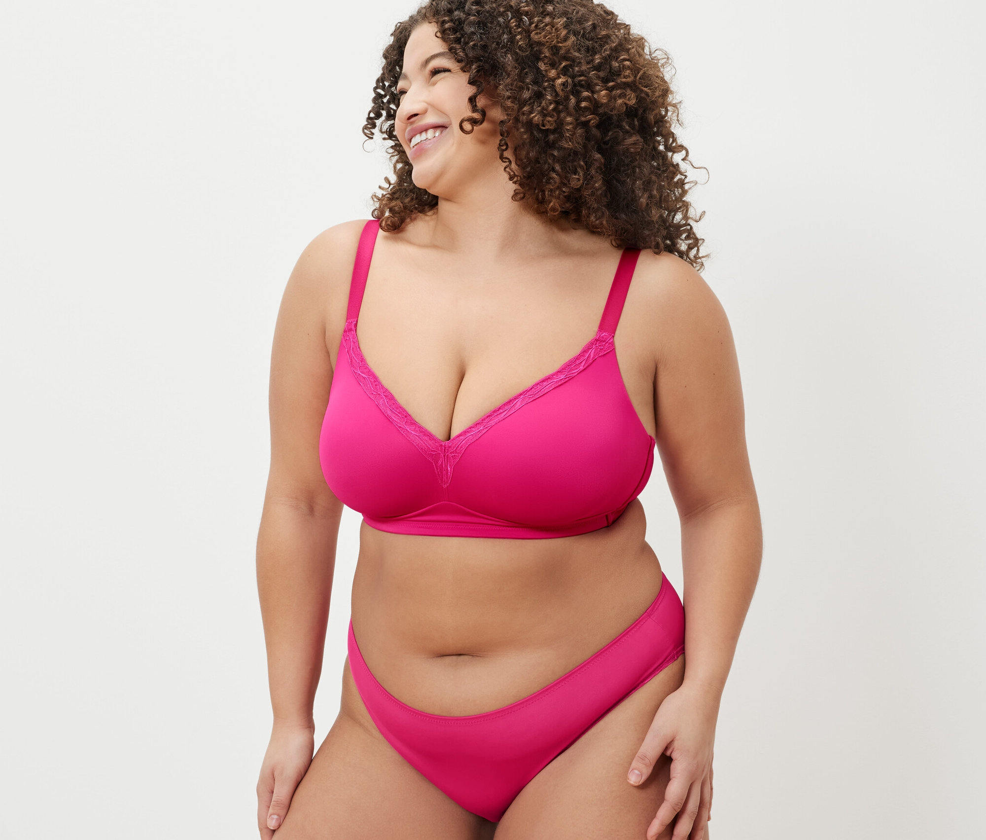 Frau mit lockigem Haar trägt einen pinkfarbenen BH und Höschen. Sie trägt einen Soft-BH und lächelt.