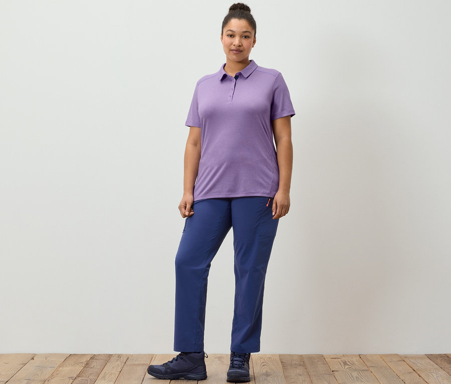 Eine Frau posiert auf einem Holzboden in dunkelblauen Outdoorhosen und einem violetten Funktionspoloshirt.