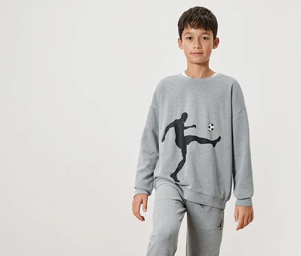 Ein Junge trägt einen grauen Pullover mit einer schwarzen Silhouette eines Fußballspielers und einem Ball darauf, sowie graue Jogginghosen.