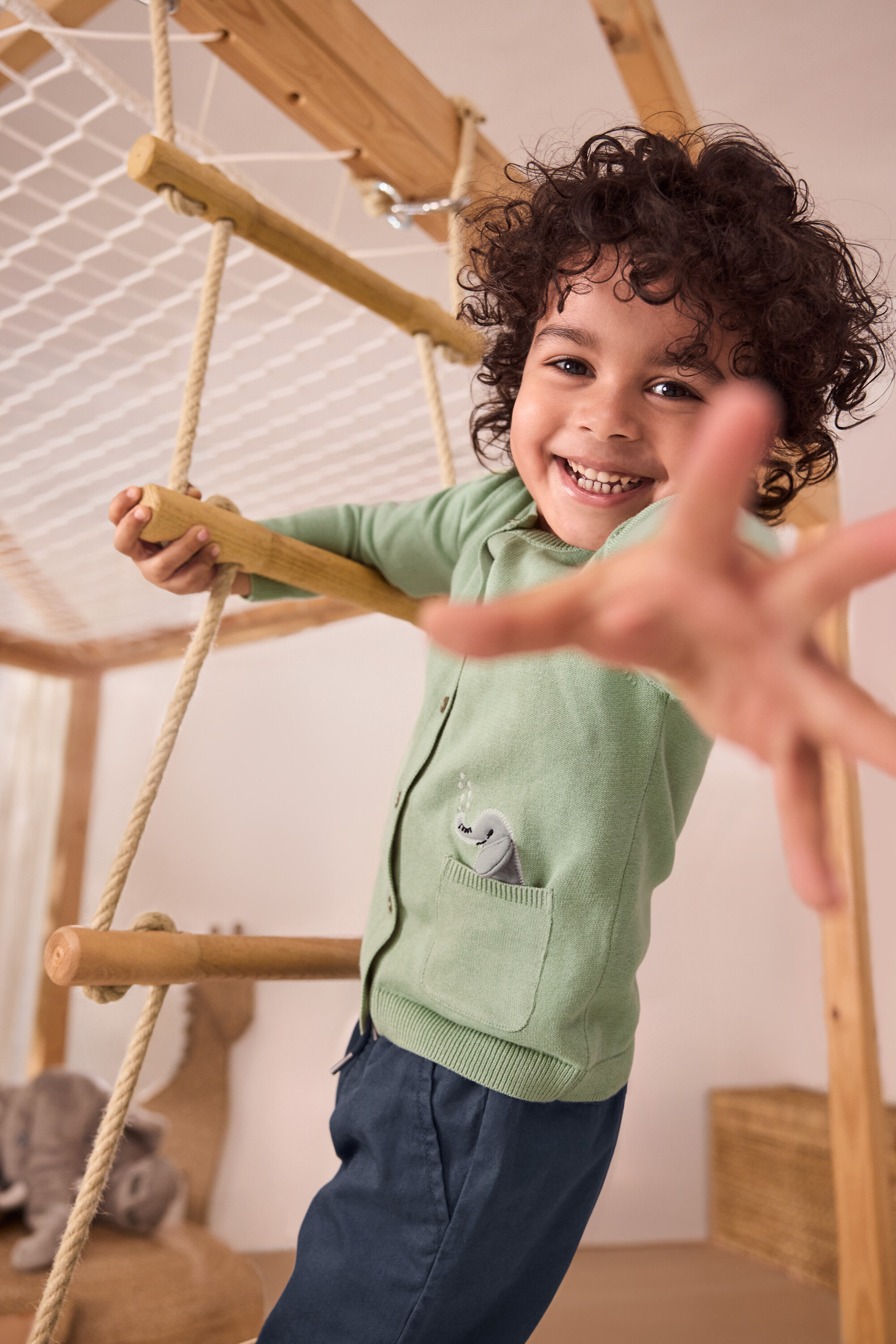 Ein lächelnder Junge klettert auf einer Strickleiter. Er trägt eine grüne Kinder-Strickjacke mit Applikation und eine blaue Kinder-Pull-on-Pants – Fit »Anton«.