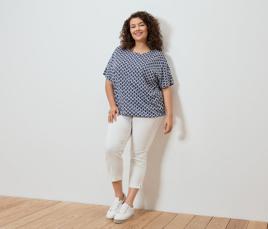 Frau mit lockigem Haar, die eine blau-weiße Bluse mit Muster, eine weiße Hose und weiße Turnschuhe trägt und vor einer weißen Wand steht.