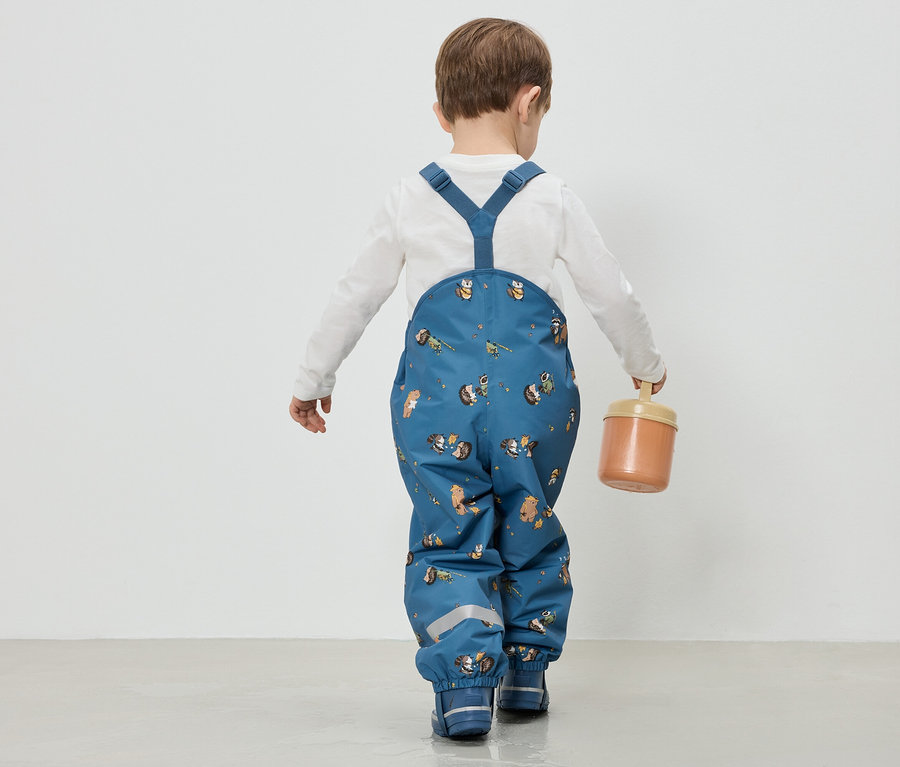 Kind von hinten in blauer Matschhose mit Tierdruck und weißen Oberteil, hält eine Dose in der Hand.