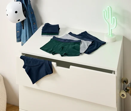 Auf einer Kommode liegen grüne, graue und blaue Boxershorts. Eine blaue Boxershort hängt aus einer Schublade. An der Wand lehnt ein Skateboard.