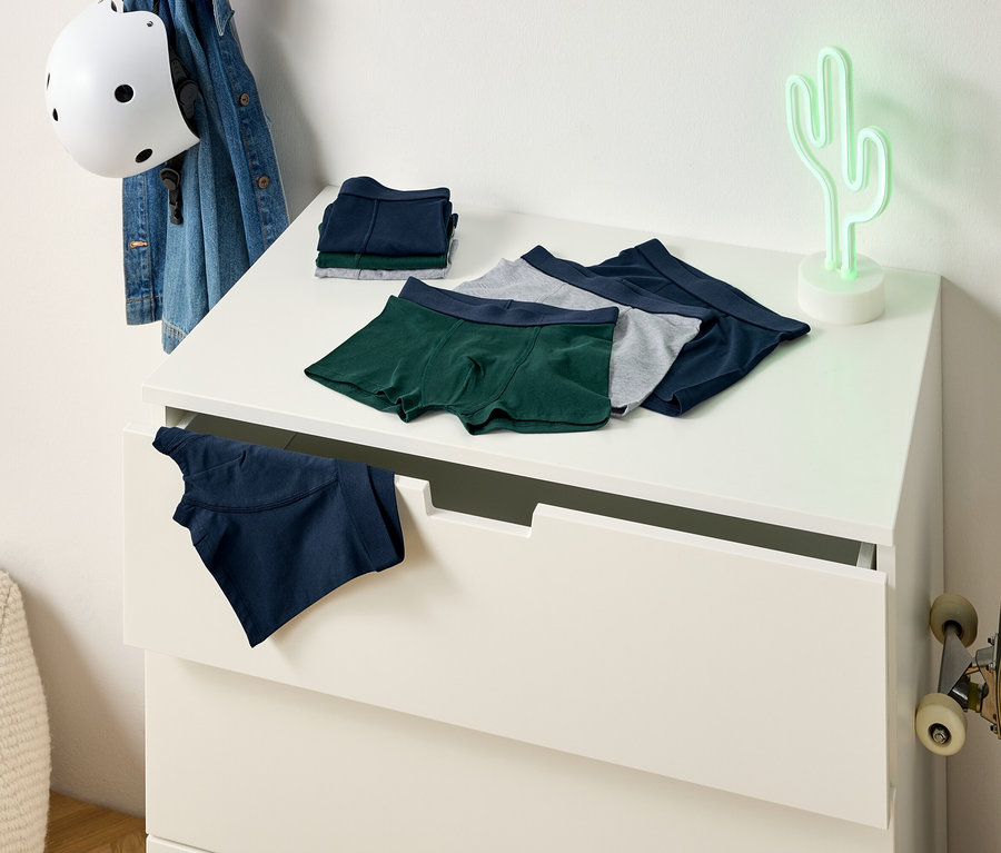 Auf einer Kommode liegen grüne, graue und blaue Boxershorts. Eine blaue Boxershort hängt aus einer Schublade. An der Wand lehnt ein Skateboard.