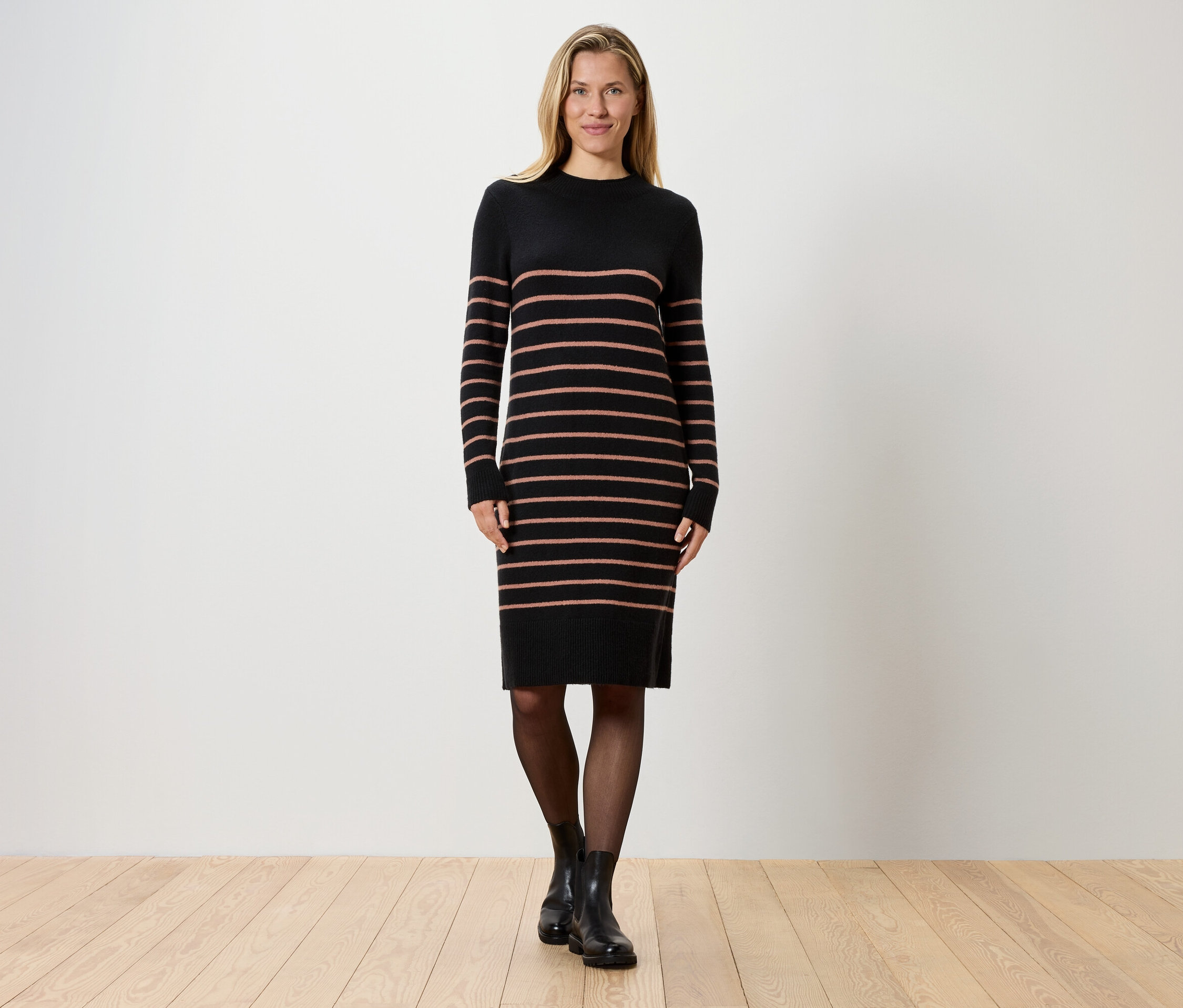 Eine blonde Frau steht vor einer weißen Wand und trägt ein gestreiftes, schwarzes Strickkleid, schwarze Strumpfhosen und schwarze Stiefel.