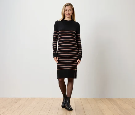 Eine blonde Frau steht vor einer weißen Wand und trägt ein gestreiftes, schwarzes Strickkleid, schwarze Strumpfhosen und schwarze Stiefel.
