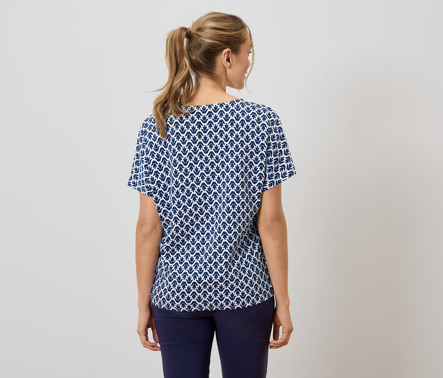 Rückansicht einer Frau mit blondem Pferdeschwanz, die eine marineblaue und weiße Kurzarmbluse mit Blattmuster und marineblaue Hosen trägt.