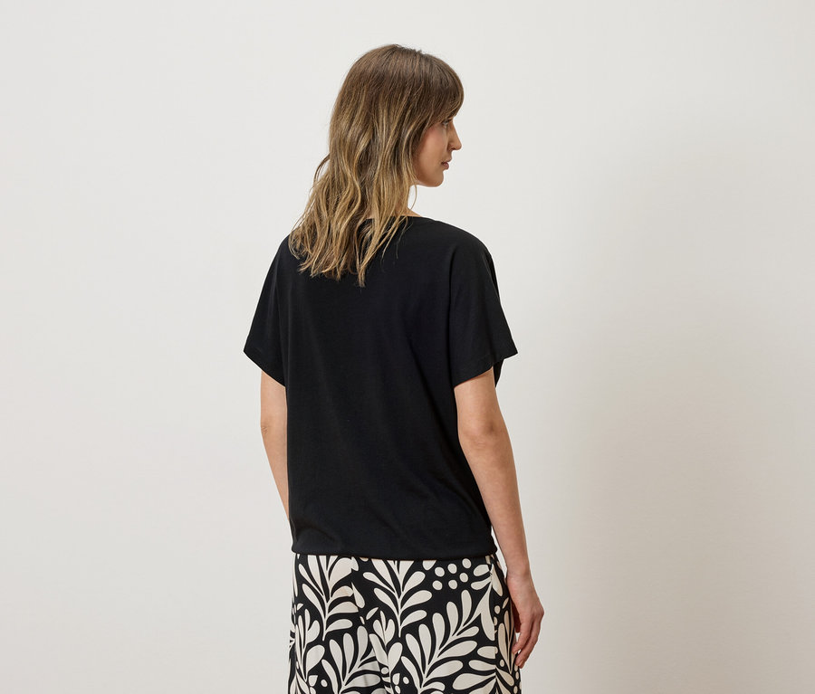 Rückansicht einer Frau mit gewelltem Haar, die ein schwarzes T-Shirt und eine Hose mit weißem Blattmuster trägt.