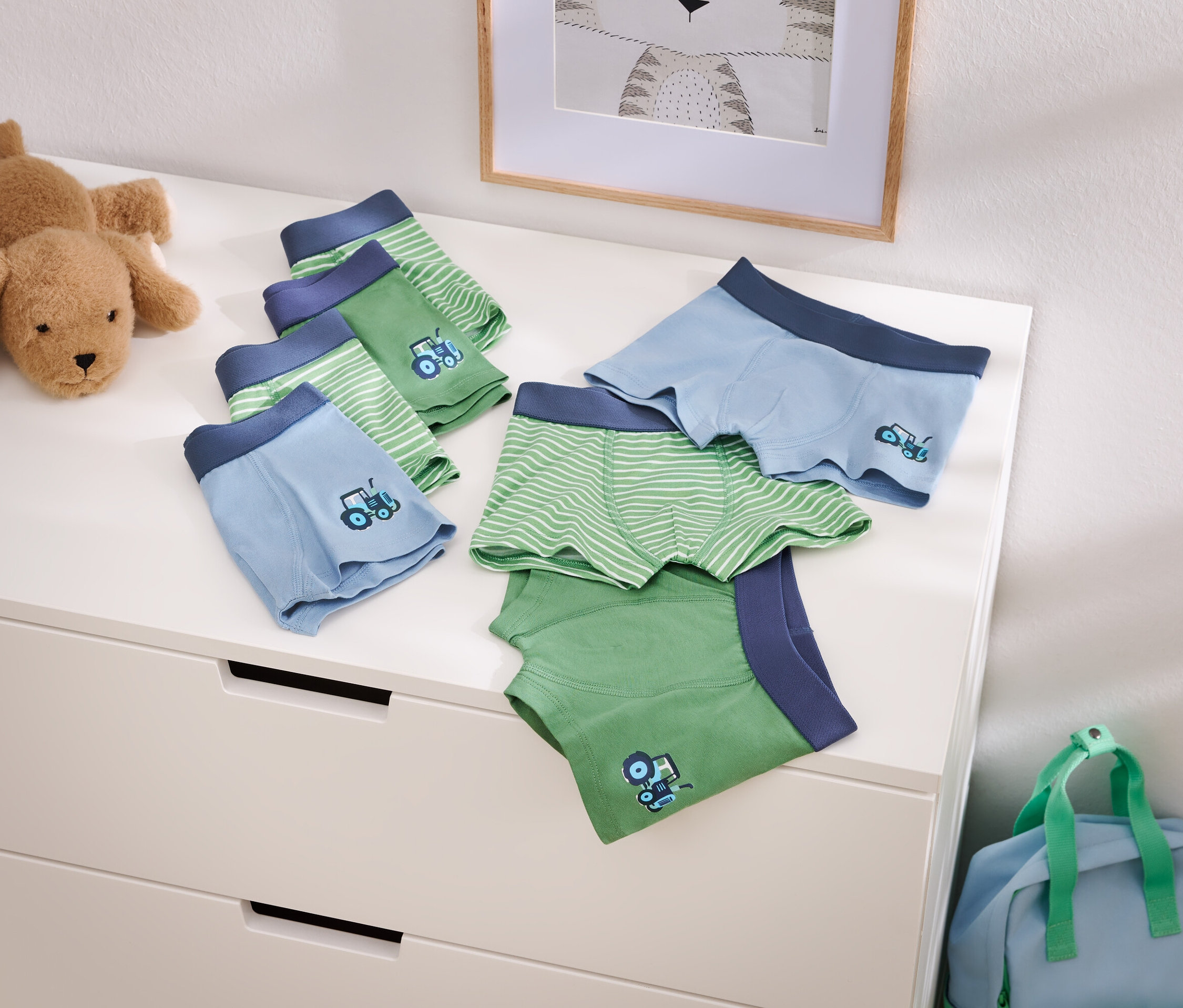 Abbildung von hellblauen und grünen Jungenunterhosen mit Traktormotiv auf einer Kommode, neben einem Stofftierhund und einer blauen Reisetasche.