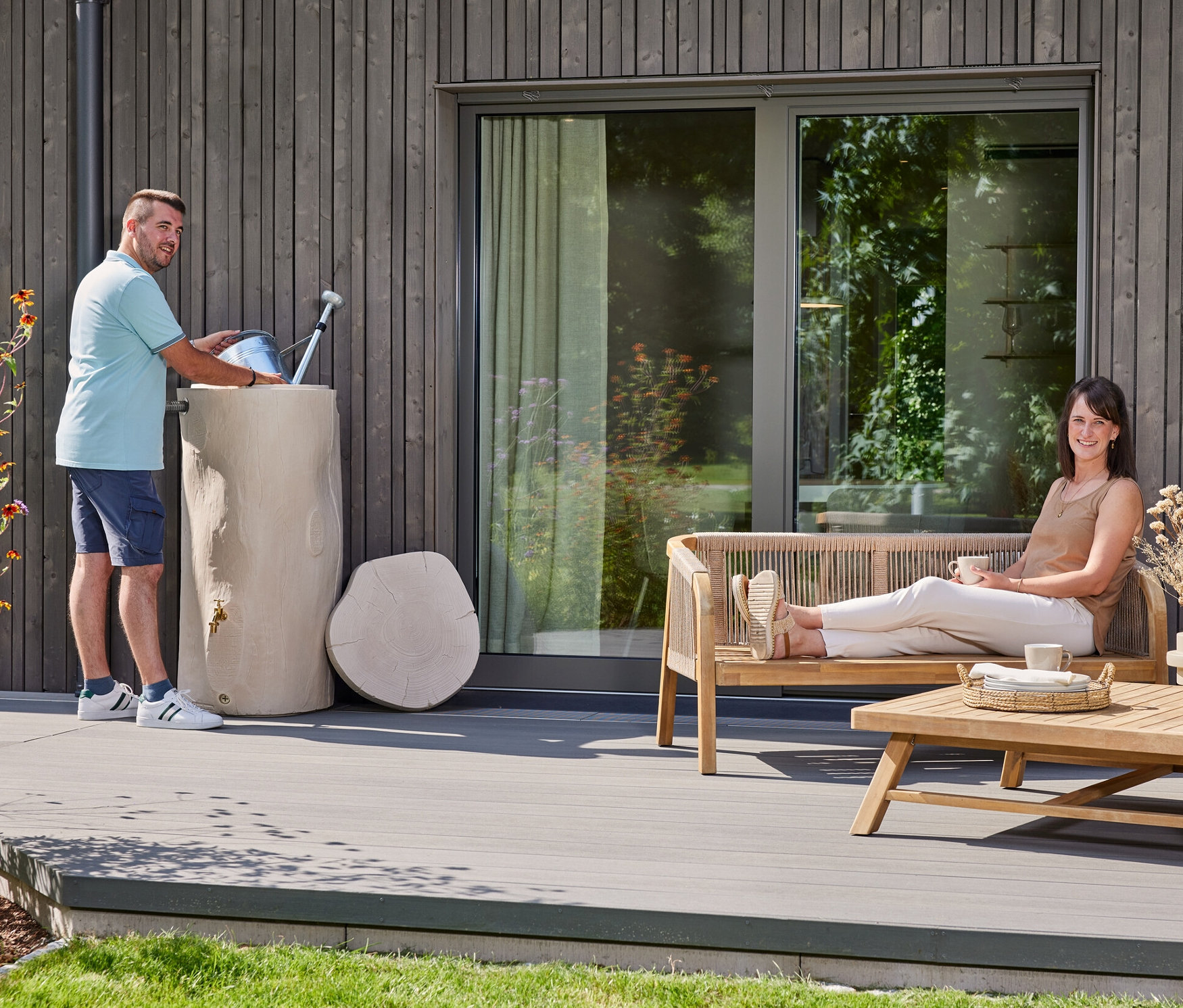 Mann gießt Wasser in eine Baumstamm-förmige Regentonne, während eine Frau auf einer Bank sitzt und eine Tasse hält.