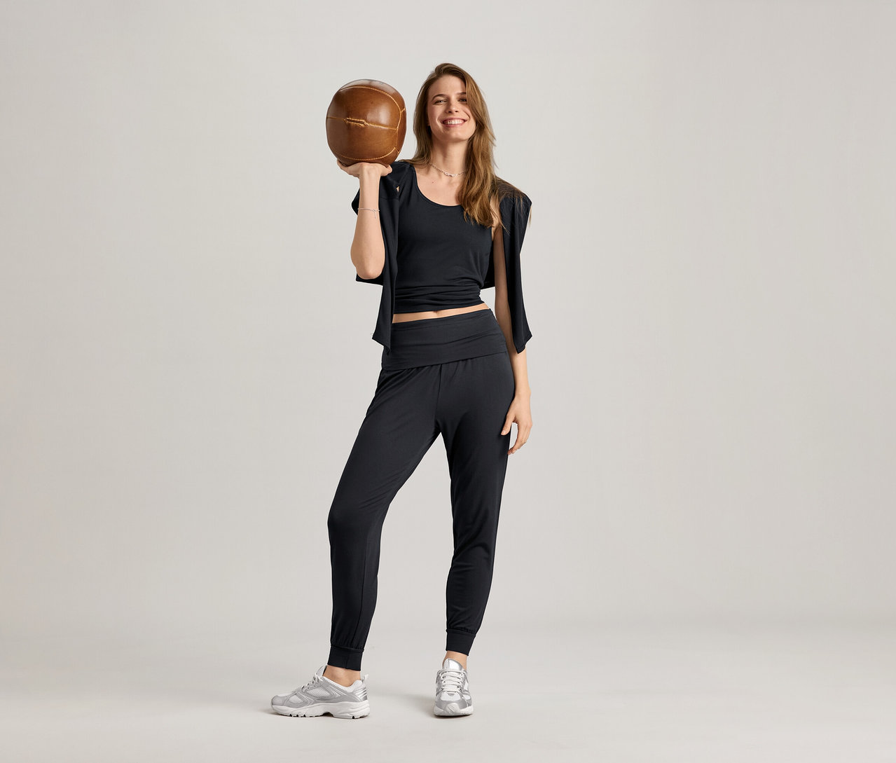 Frau im schwarzen 2-in-1-Sportshirt, mit Sporthose und Turnschuhen, hält einen braunen Ball hoch.