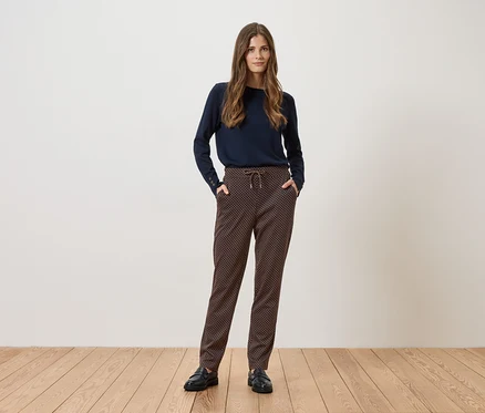 Eine Frau steht vor einer weißen Wand und trägt einen marineblauen Pullover, gemusterte Hose und schwarze Schuhe.