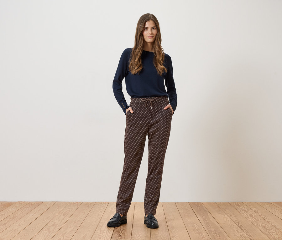 Eine Frau steht vor einer weißen Wand und trägt einen marineblauen Pullover, gemusterte Hose und schwarze Schuhe.