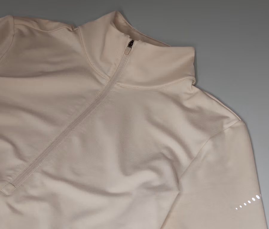 Beige Sportjacke mit Reißverschluss und hohem Kragen liegt auf grauem Hintergrund.